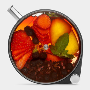 A glass teapot filled with tea and various ingredients, including sliced strawberries, blueberries, a lemon wedge, mint leaves, and tea leaves.